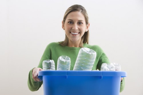 A young woman with shoulder-length light brown hair, smiling and wearing a bright green sweater, holding a blue plastic recycling bin filled with clear plastic bottles of various sizes, some with ridged textures, positioned in front of her against a plain light grey background. The bottles are arranged upright in the bin, with the larger bottles towards the back and smaller ones at the front, indicating a collection for recycling or rubbish disposal. The scene suggests an environmentally conscious approach, aligned with waste management services such as those offered by Commercial Waste Colindale, serving areas around NW London, including Colindale. The lighting is soft and evenly distributed, highlighting the clarity of the plastic bottles and the cheerful expression of the individual, reinforcing a professional and approachable image of rubbish collection and recycling efforts in a domestic or small commercial setting.