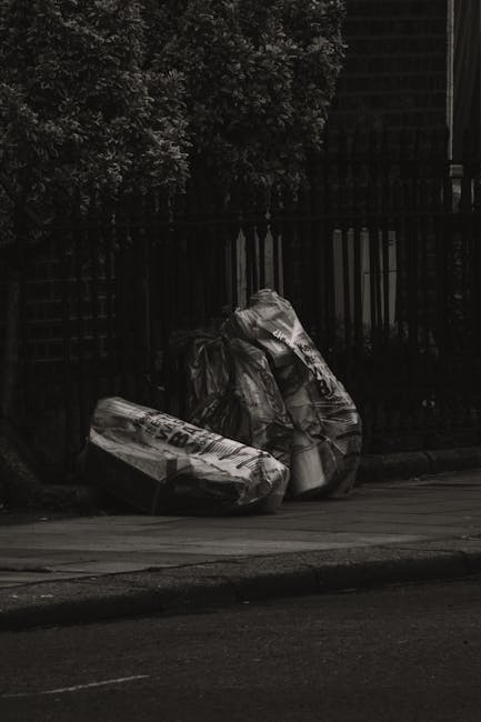 Two large black plastic garbage bags and a cardboard box, all filled and tied at the top, are placed on the pavement beside a black metal fence. The bags appear to contain household waste or debris, e